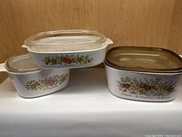 Three Corning bakeware casserole dishes stacked and shown with lids, displaying varying lid styles and sizes, set on wood background.