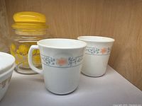 Two white Pyrex coffee mugs with orange and blue-gray floral band pattern alongside a yellow-lidded glass jar with dandelion print.