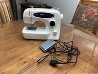 Front right side view of white Brother XL-2230 sewing machine with purple stitch selector panel and visible sewing area. The power cord and grey foot pedal are placed in front on the wooden table.