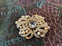 Close-up of the vintage gold tone Swarovski brooch showing the clear cushion center crystal, surrounding smaller crystals, cream-colored pearls, and gold metal loops on fabric background.