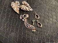 All jewelry pieces on a black textured surface showing large leaf-shaped gold tone earrings, smaller engraved earrings, and peacock brooch