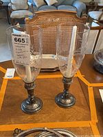 Pair of weighted sterling silver candlestick holders with tall etched glass tops containing white taper candles, sitting on a wood table with a cane back chair in background.