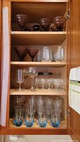 Full cabinet with 4 shelves showing various glassware including blue tone glasses, stemware, and purple glass candle holders