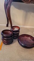 Stack of eight small wooden bowls in dark polished wood finish alongside one larger serving bowl of the same finish, placed on carpeted floor near wooden furniture leg.