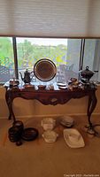 Full view of the wooden hall table with decorative carvings shown by a window, surrounded by various household items placed on and under the table, which are not included in the lot.