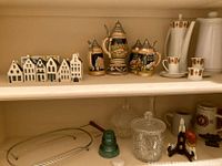 Shelf view showing four Dutch miniature houses, four German beer steins, white ceramic coffee set, clear glass jar, green glass insulator and small ceramic building
