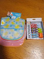 Photo of three foldable silicone baby bibs with yellow duck patterns and a boxed Boon Modware toddler utensil set in colorful handles