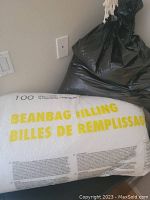 Large clear plastic bag of bean bag filling with yellow text, plastic bag partially opened with filler transferred to black plastic bag beside it