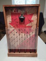 Front view of the wooden Plinko game board with red star and bottle opener mounted on the backboard, clear plastic guard, and multiple nails arranged on the pegboard.