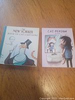 Two cat-themed books on a wooden surface: The New Yorker Book of Cat Cartoons with a man in top hat on cover and Cat Person by Jieun Kim with cartoon girl and cat illustration.