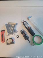 Photo showing hardware and tools including tape measure, utility knives, angle square, flashlight, screwdriver, painters tape, and zip ties arranged on a white surface.
