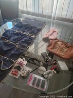Overview of lot contents on glass table including hanging shoe organizer, brown studded handbag, various hair accessories, belts, costume jewelry, headbands
