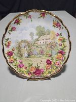 Front view of the Old Country Roses collector's plate showing a cottage and garden scene, with a decorative border of pink, red, and yellow roses and gold trim.