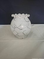 Front view of the white ceramic vase with black abstract lines and wavy ruffled rim, shown on white cloth background.