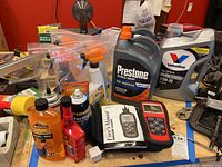 Photo showing automotive code scanner, synthetic motor oil, Prestone coolant, multiple cleaning supplies and automotive fluids on a table with a busy background.
