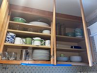 Kitchen cabinet open displaying Studio Nova plates, mugs, bowls, glass pitchers, casserole dishes, and serving bowls arranged on shelves.