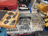 Overview of workbench with wooden tap and die set next to plastic drill bit case and other tools scattered in background.