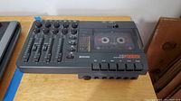Top-down view of the Fostex X-18 multitracker showing four channel sliders and rotary knobs, cassette tape inserted in transparent compartment, multiple transport buttons on front.
