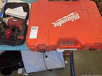 Closed Milwaukee red plastic carrying case with Skil hammer drill in a zipped carrying bag to the left on the table.