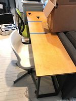 Sofa table with light wood top and black metal frame, showing surface scratches and blue tape pieces, accompanied by a white plastic chair with metal rolling base and green mesh office chair with rolling base in background.