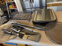 Overview of grilling utensils including tongs, spatula, fork, basting brush with wire racks and a round stone or ceramic hot plate