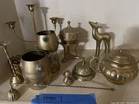 Photo showing assortment of brass items including candlesticks, cups with stones, covered containers, teapot, deer figurine, hand bell, two small knobs, and stirrer rod