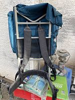 Back view of blue Camp Trails hiking backpack mounted on aluminum frame with padded waist belt and shoulder straps, resting against outdoor surface with some camping gear visible near lower part.