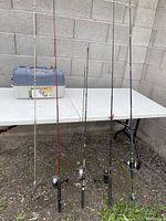 Full view of 5 fishing rods with reels and tackle box on table outdoors