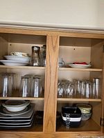 Wooden kitchen cabinet shelves showing glass drinking glasses, plates, bowls, and ceramic containers.