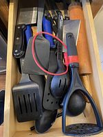 Drawer with assorted black and blue cooking utensils including spatulas, ladle, potato masher, wooden rolling pin.