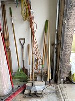 Photo showing yard tools including rakes, shovel, broom, axe, cultivator tools placed against a wall in a garage.