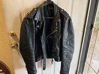 Front view of black leather men's riding jacket hanging on hanger showing zippered front and belted waist.