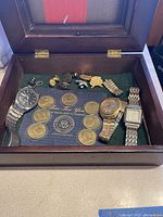 Wooden box open displaying three metal band men's wristwatches, brass coins, and small rings