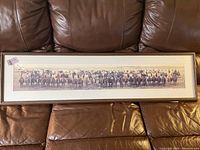 Framed panoramic photograph of a line of cowboys on horseback, laid out on a leather sofa, measuring 51x14 inches