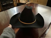 Black men's western hat shown on table, gold rope band, wide brim.