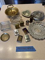 Photo showing all ashtrays and lighters together on table, including metallic and glass ashtrays and eight flip-top lighters