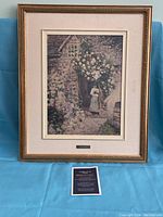 Framed replica artwork showing a stone cottage covered with white flowers and a woman at the door. The piece includes an informational card about the reproduction company.