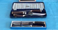 Two MCM cutlery sets displayed in cases; one in a blue velvet-lined box, the other in a blue cardboard box.