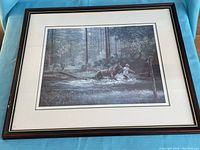 Framed print shows man leading horse through stream in forest setting with visible frame damage.