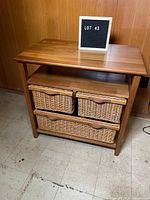 Wooden shelving unit with a natural finish, showing top and middle shelves with two small wicker basket drawers and one large wicker basket drawer on the bottom shelf, placed on a tiled floor against a wooden-paneled wall.
