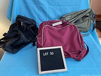 Three small duffle bags, black, maroon with white piping, and olive green, arranged against a blue background with a black letter board marked 'LOT 30'.