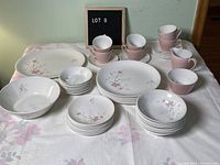 Photo showing full set of dishes including serving platter, serving bowl, cups, saucers, plates, and bowls with pink floral pattern on white base and solid pink cups.