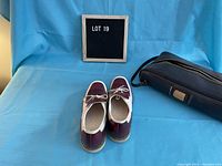 Front and side view of vintage burgundy and white women's bowling shoes with carrying bag in background on blue backdrop