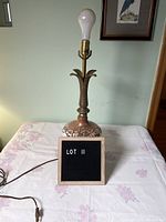 Full view of vintage ornate table lamp showing base, stem, socket with bulb, on floral tablecloth against wall with framed art.