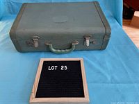 Green hardcover vintage suitcase shown closed with visible wear and metal clasps, placed on blue background, includes lot number board.