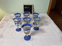 Seven blue-based wine goblets with blue rims and hand painted floral blue and yellow designs on the clear glass body arranged on a table.