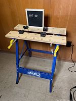 Front view of the portable workbench showing blue metal legs, wooden top with measurement markings, yellow side clamps, and black top clamps.