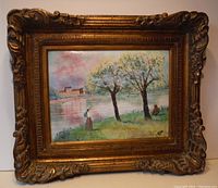 Frontal photo of enamel on copper landscape painting with ornate gold frame showing two trees, two figures, river, and a building across the water.