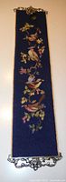 Full view of a vertical blue tapestry with colorful embroidered birds and flowers, mounted on an ornate brass frame with a small bell at the top center.
