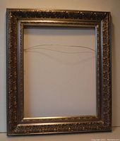Front view of an ornate empty picture frame with gold-toned decorative border and hanging wire on the back.